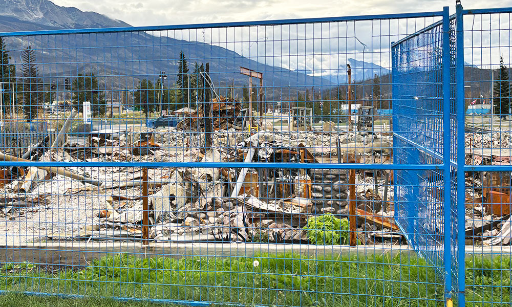 A chain-link fence with blue framing runs across the foreground, behind which lies a scene of devastation. The remnants of a burned-down house are visible, with charred debris and twisted metal scattered across the lot. Mountains and trees are visible in the background under a cloudy sky.