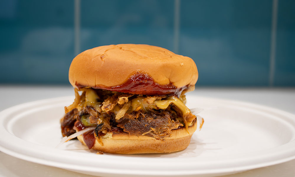 a shredded meat burger on a paper plate. there's barbecue sauce on the side of the bun and melted cheese under it