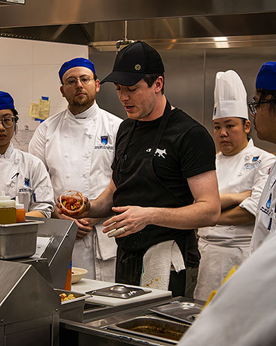 a chef in a black ballcap and t-shirt shows a group of students in chefs whites a cooking technique in a commercial kitchen