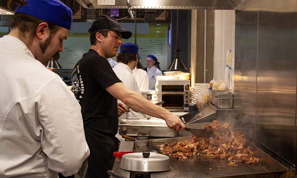 a chef in a black ballcap and t-shirt turns shredded meat on a commercial grill