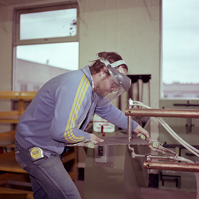 A person wearing a protective face shield and blue jacket is working on a sheet metal piece using a specialized cutting or bending machine in an indoor workshop.
