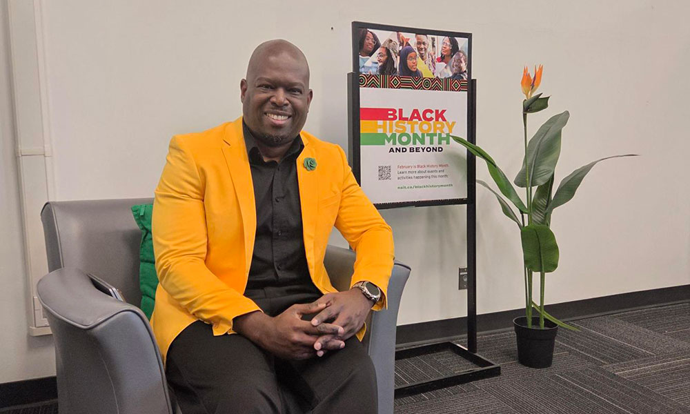 A person seated in a chair wearing a bright yellow jacket and dark clothing, positioned beside a “Black History Month and Beyond” sign and a potted plant.