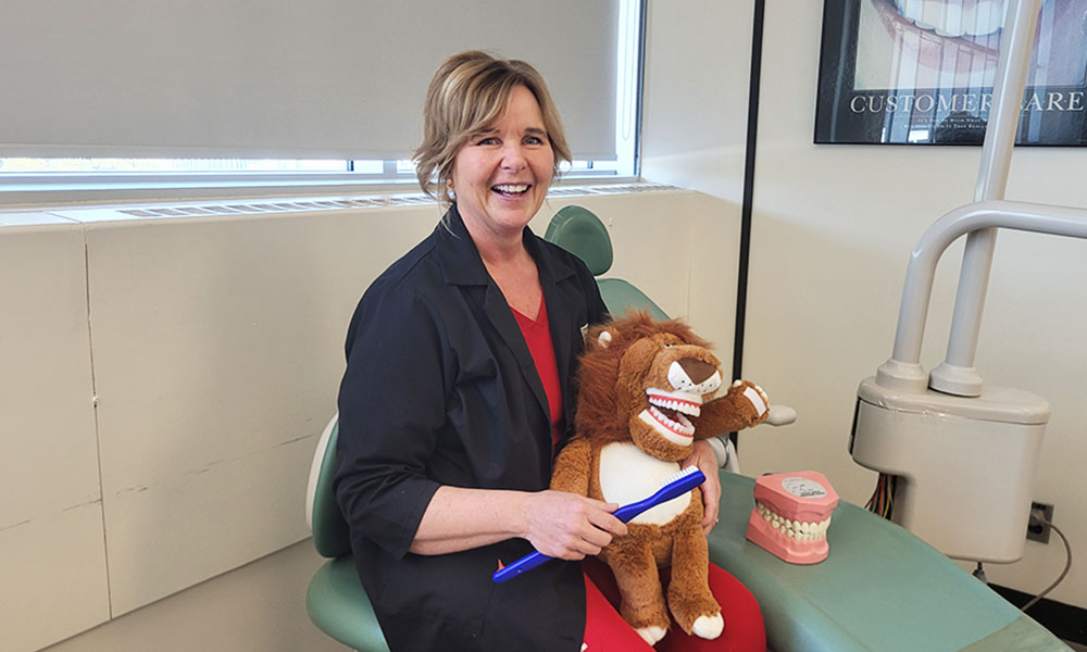 NAIT Dental Assisting instructor and grad Sherry Sand Adult seated in a dental clinic holding a plush toy and toothbrush beside a dental chair.
