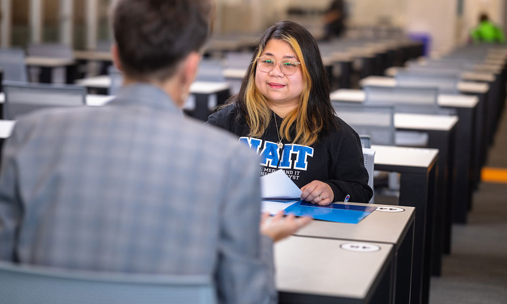 Two people sit across from each other at a table in a large room with rows of desks; one has a NAIT sweatshirt and a blue folder.