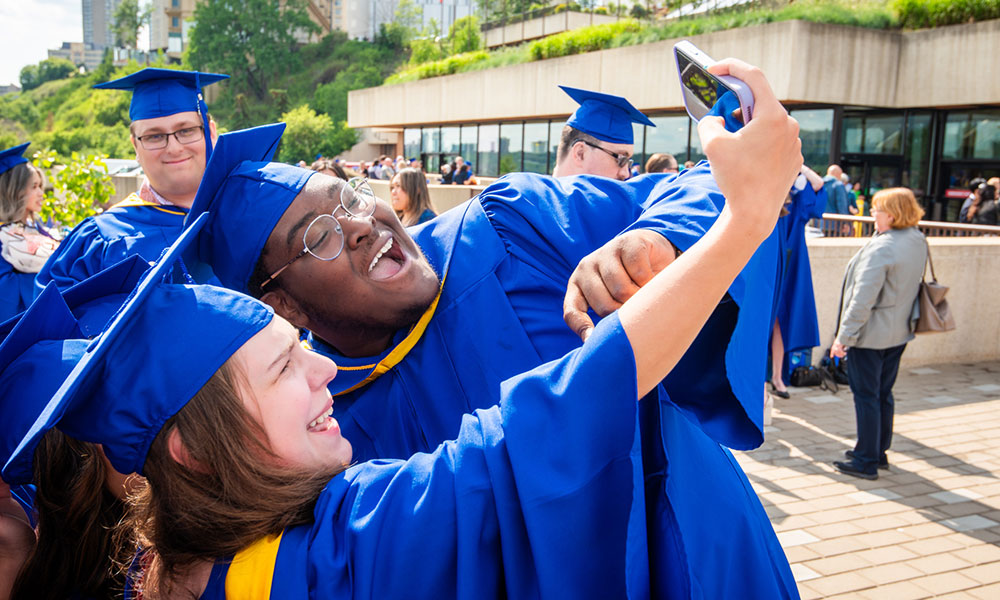 Group of people in blue graduation gowns and caps taking a selfie outdoors, with greenery, other people, and a building with large windows in the background.
