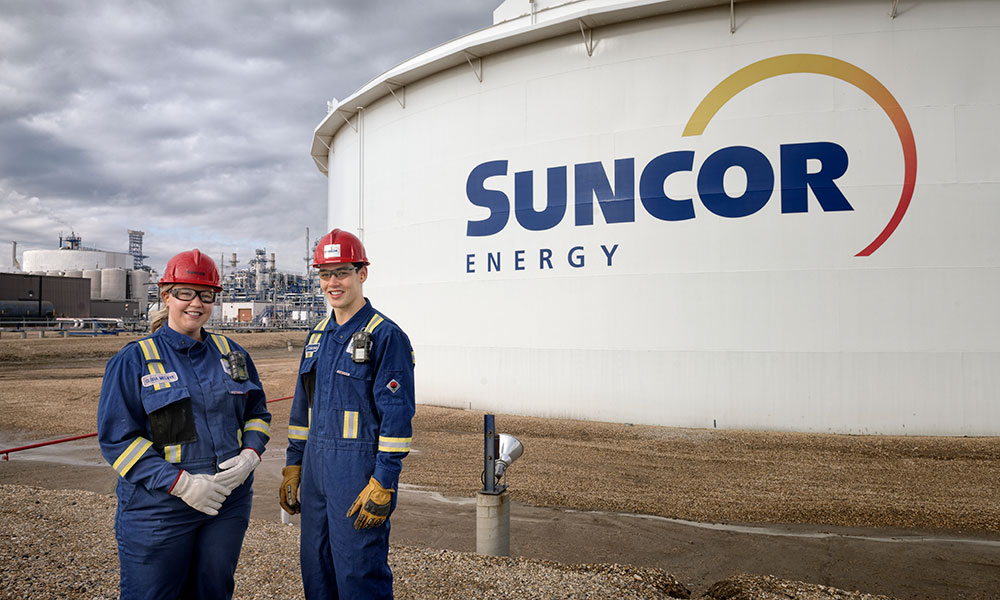 Two workers in protective gear stand at an industrial site in front of a large storage tank labeled “Suncor Energy,” with refinery structures in the background.
