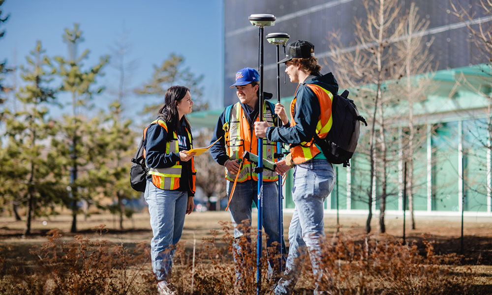 Three individuals in safety vests conducting a surveying activity outdoors, with trees and a building in the background. One person holds a surveying instrument mounted on a pole.