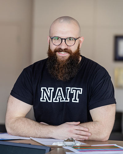 Smiling bald man with glasses and a full beard, wearing a black NAIT T‑shirt, seated at a desk with papers.