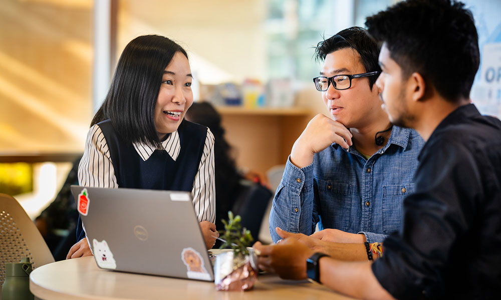Three people sitting at a round table with a laptop, engaged in conversation in a bright indoor setting.