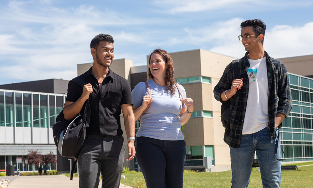 Three individuals walking outdoors on a campus with modern buildings in the background, carrying backpacks and engaged in conversation.