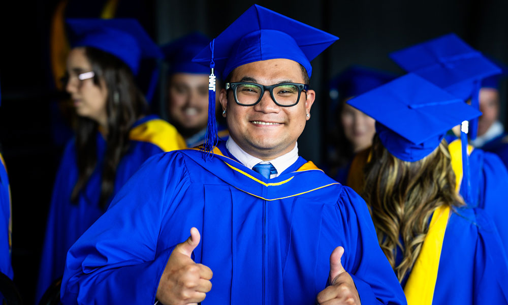 Group of people in blue graduation gowns and caps with yellow accents. One person in the foreground is giving two thumbs up."