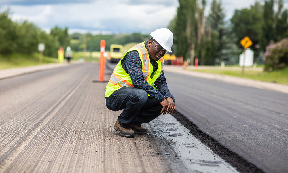 a man in a white hard hat and yellow safety vest crouches to examine freshly laid pavement