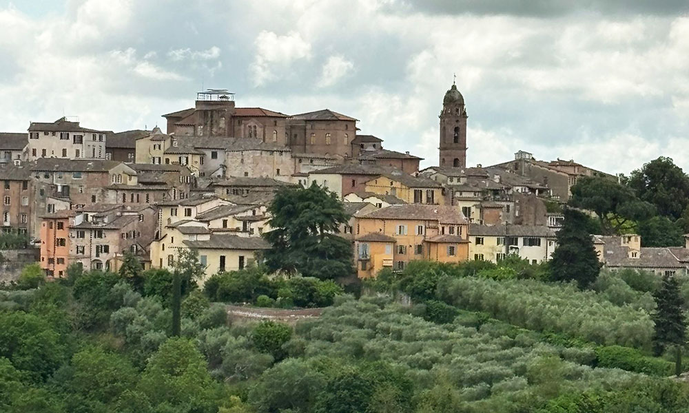 Picturesque view of a hilltop town with colorful buildings in beige, yellow, and orange, surrounded by lush greenery. A tall bell tower stands prominently on the right under a partly cloudy sky.