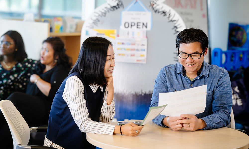 Two people sitting at a round table in an indoor setting, holding papers and discussing information.
