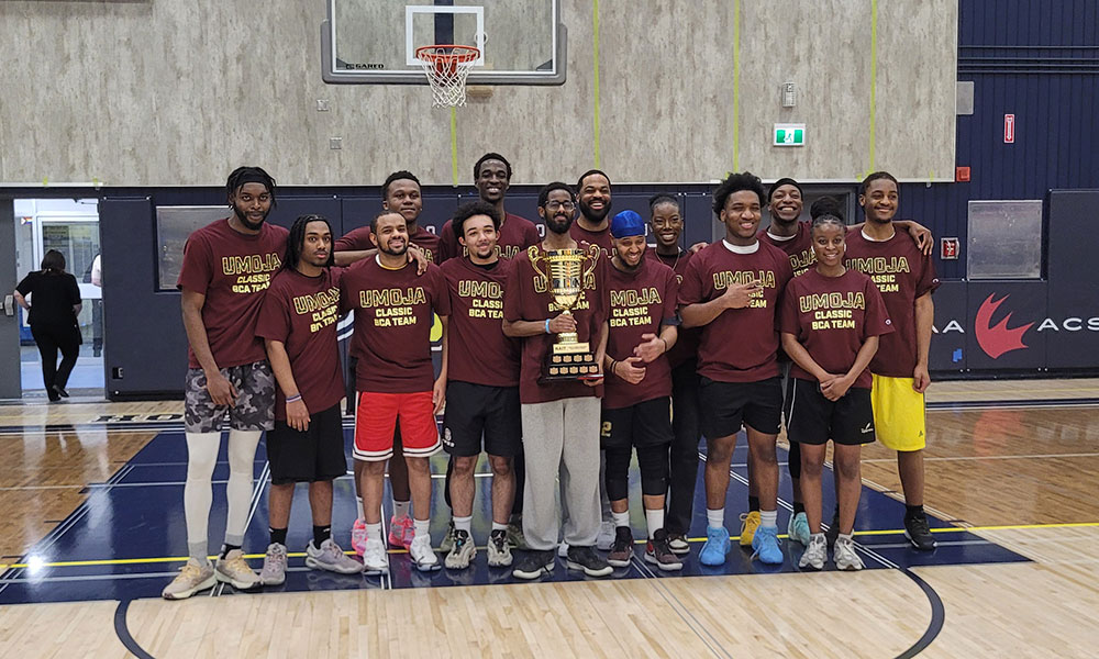 A basketball team wearing matching “UMOJA Classic BCA Team” shirts stands together on the court, gathered around a large gold trophy.
