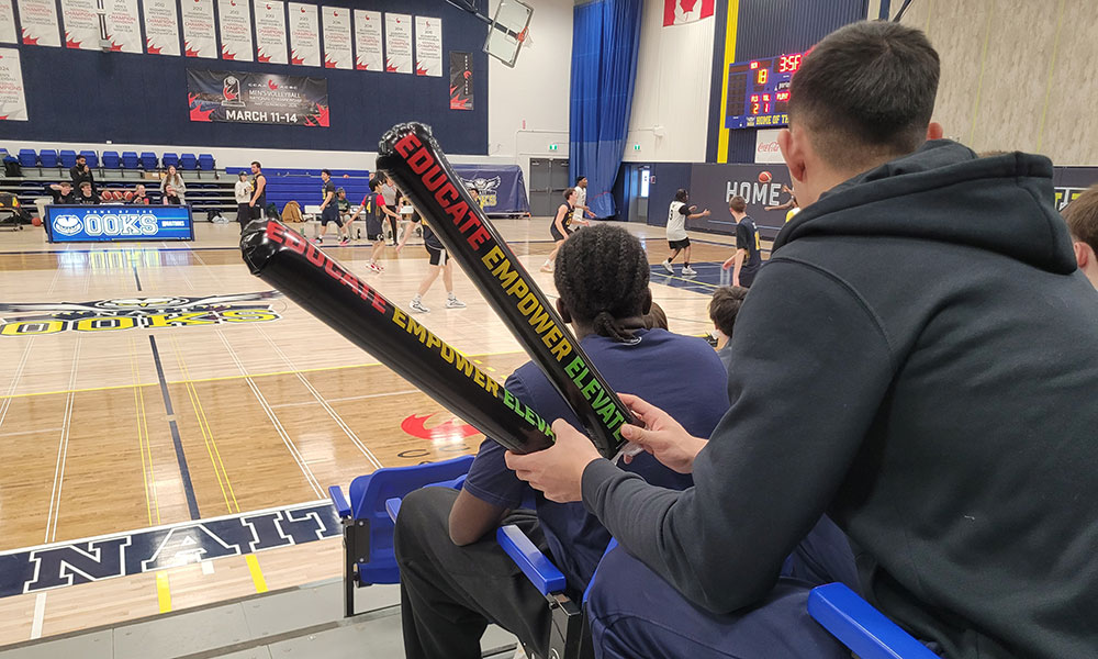A spectator sits in the stands holding two colorful cheer sticks that read “EDUCATE • EMPOWER • ELEVATE” while watching a basketball game underway on the court.