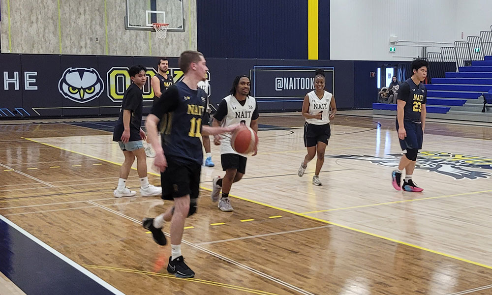 Players from two NAIT basketball teams scrimmage on the court as one player dribbles the ball up toward the key.