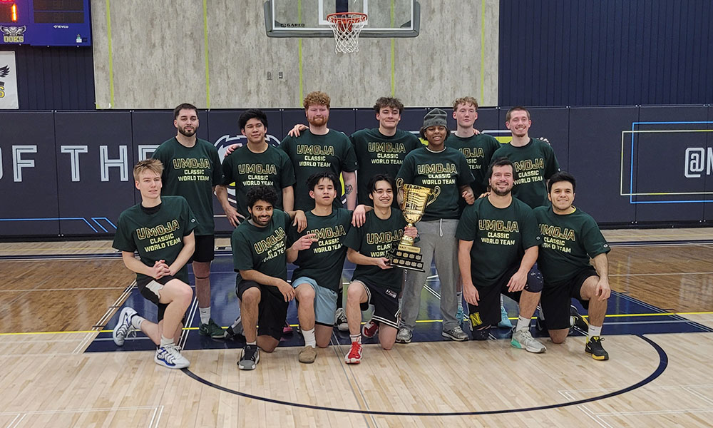 A basketball team wearing matching “UMOJA Classic World Team” shirts poses together on the court around a large gold trophy.