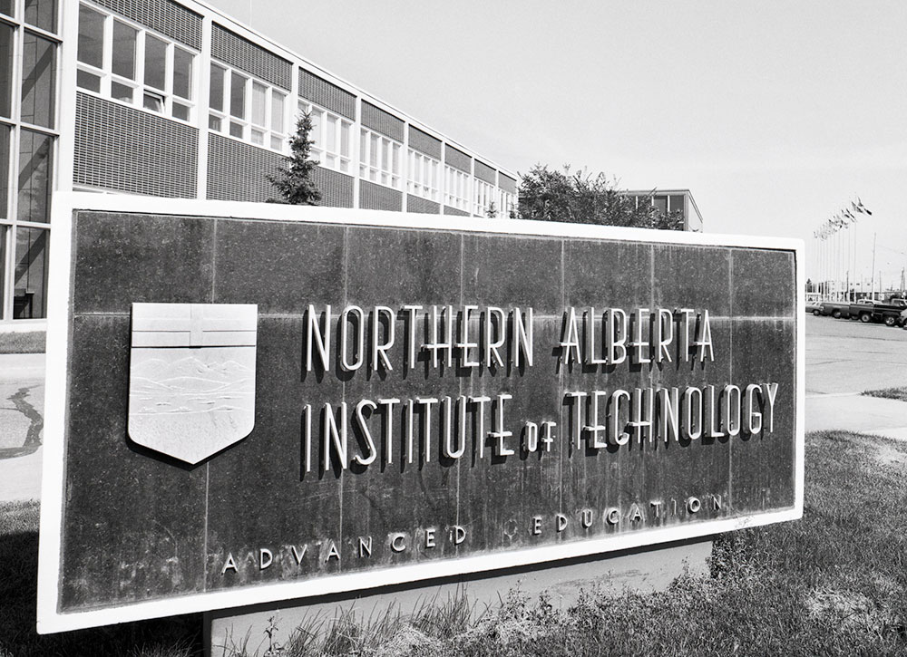 Black-and-white photo of a large stone sign reading “Northern Alberta Institute of Technology – Advanced Education” with a crest, in front of a mid-century modern campus building with long rows of windows.