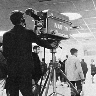 Person operating a large vintage television camera labeled ‘NAIT-TV’ on a tripod in an indoor public space, with several people walking in the background.