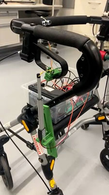 Close-up of a walker modified with electronic components, wiring, and a clear plastic box containing circuit boards. Green brackets and sensors are attached to the frame, suggesting an engineering or assistive technology project.