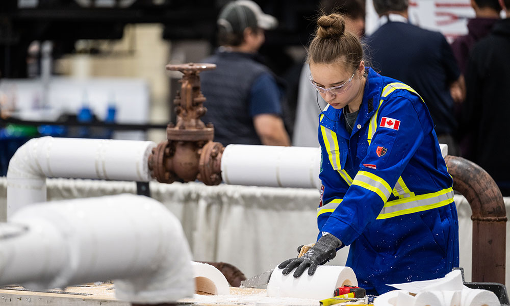 A person in a blue uniform with reflective yellow stripes and a Canadian flag patch on the sleeve is working with white pipes and fittings in an industrial or construction setting. Other individuals are visible in the blurred background.