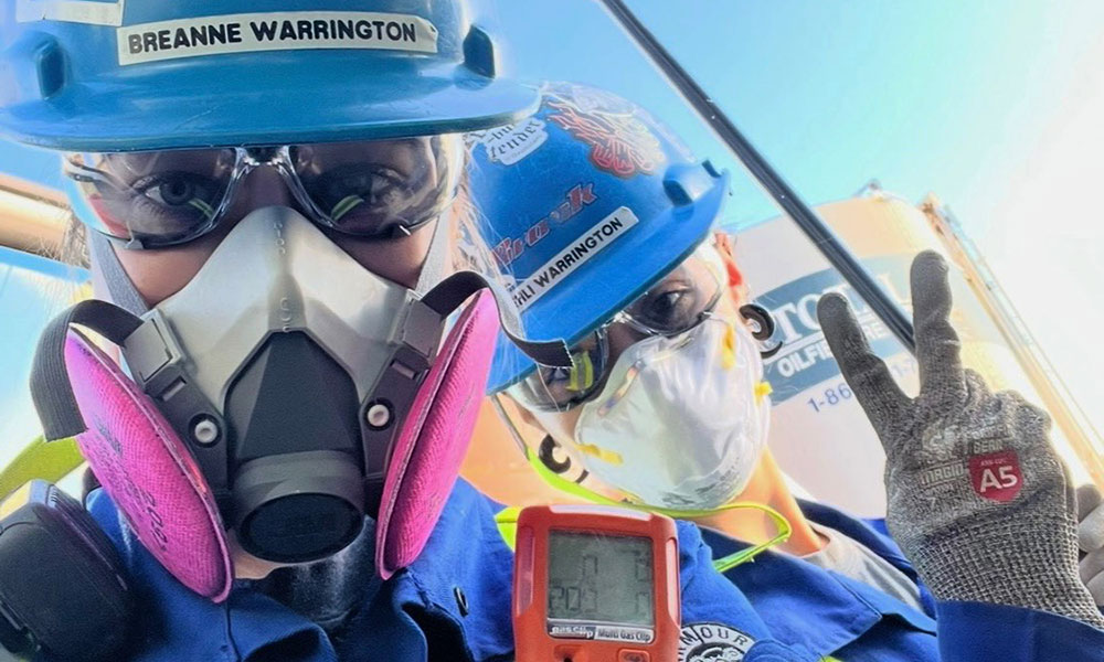 Two individuals wearing blue hard hats, masks, and gloves. The person on the left has a blurred face and a name tag reading 'BREANNE WARRINGTON.' The person on the right, with a name tag reading 'KEHLI WARRINGTON,' is holding up a peace sign and a device displaying numbers.