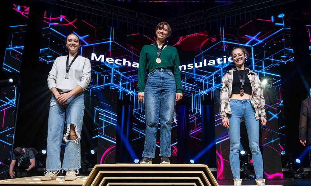 Three individuals stand on a podium wearing medals around their necks. Behind them is a digital display with geometric patterns and partially visible text reading 'Mechanical insulation.'