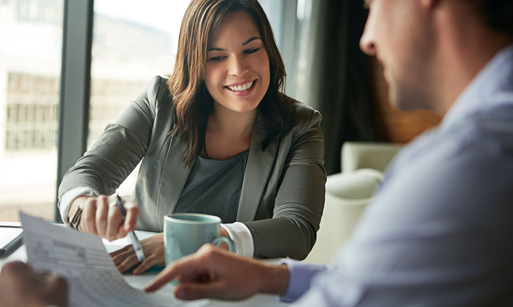 A woman smiling as she reviews a document with a man at a table, with a coffee mug between them.