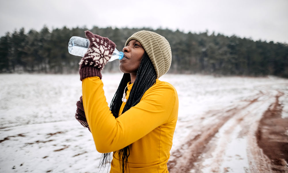 woman in yellow shirt and beige toque drinking water outdoors after a winter workout