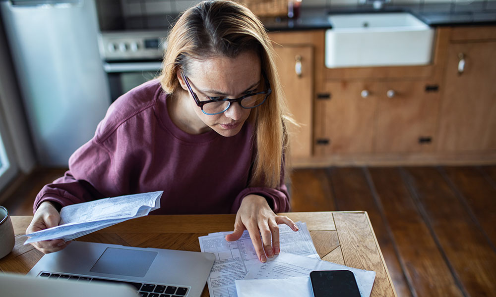 A woman at a kitchen table reviewing paperwork with a laptop and phone in front of her.