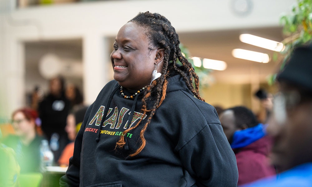 A person wearing a black NAIT hoodie stands indoors in a busy communal space with other people in the background.
