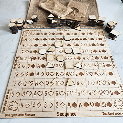 A wooden game board for Sequence is shown with engraved card symbols arranged in a grid. Several round and triangular wooden markers are placed on the board, and more markers are scattered around along with burlap bags.
