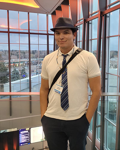 A person wearing a fedora, white shirt, and striped tie stands in a modern glass-walled atrium with city views in the background.