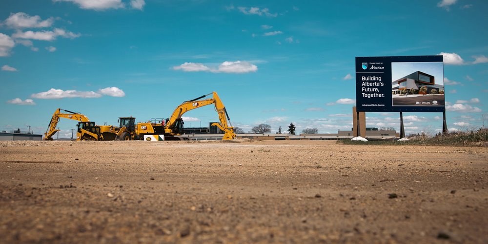 Wide-angle view of an active construction site with yellow excavators and machinery on a dirt lot, alongside a roadside sign reading “Building Alberta’s Future, Together” under a bright blue sky.