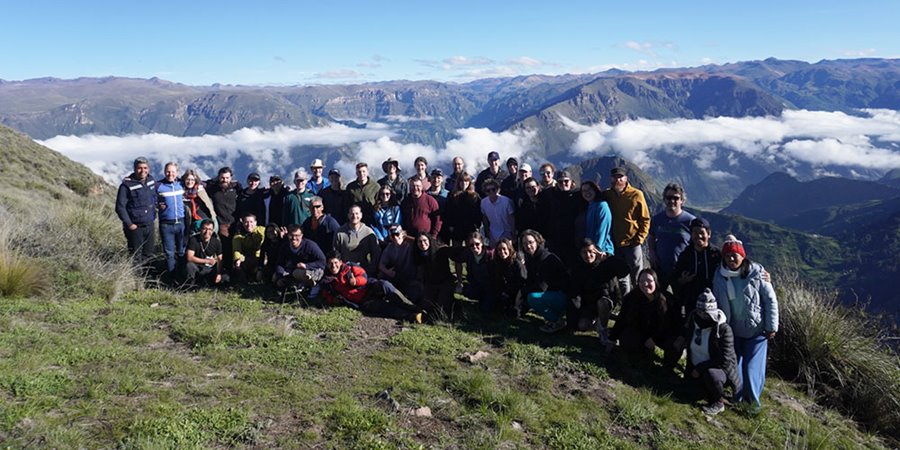 A large group of people stand and crouch together for a group photo on a grassy hillside, with a wide mountain valley and clouds visible in the distance.