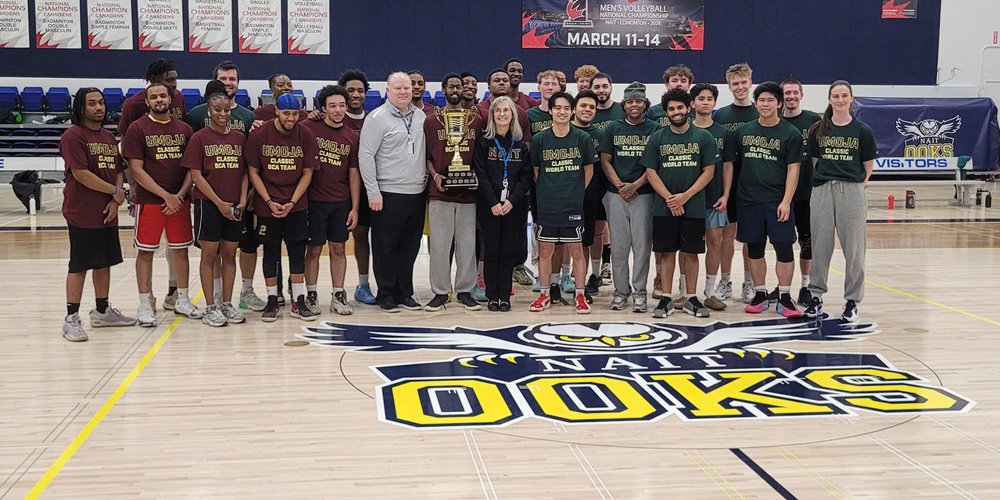 Group photo of a basketball team and coaches standing on a gym court with a championship trophy, NAIT Ooks logo on the floor, and Umoja Classic banners in the background.