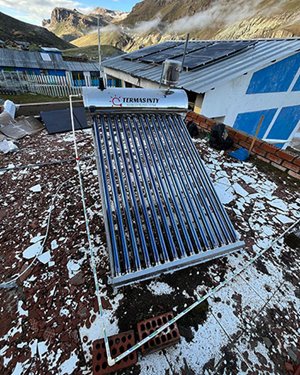 A long, silver solar water heater with blue tubes, labeled "TERMAS INTY," rests on a rough, damaged rooftop with scattered debris, against a backdrop of mountains and a village.