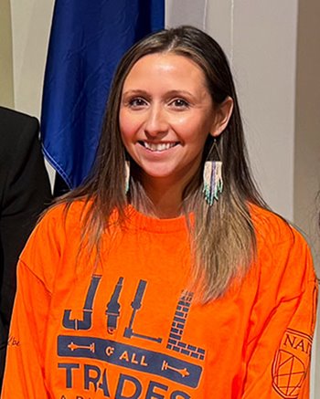A person is wearing a bright orange long-sleeve shirt with the text “Jill of All Trades” and tool graphics on the front. The individual has long, straight hair and is wearing distinctive, dangling beaded earrings in light colors.