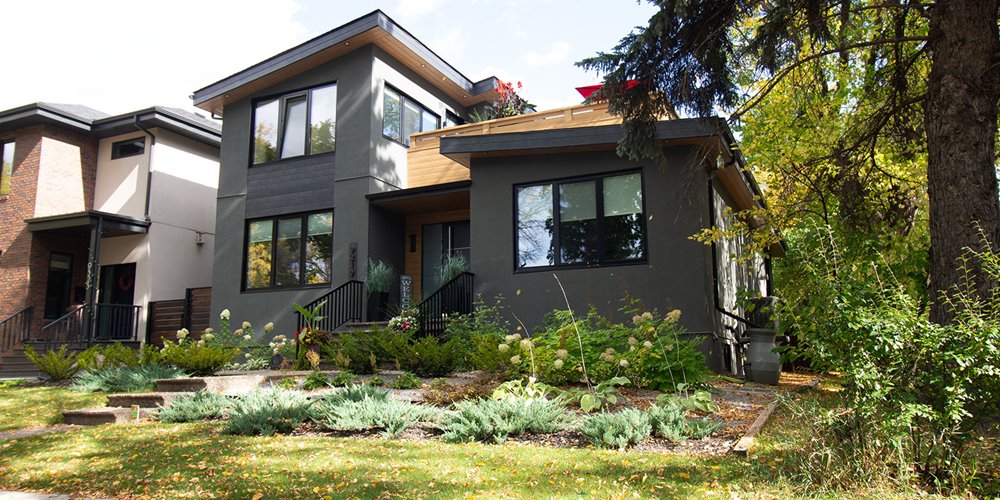 A modern two-story house in dark gray with black windows sits among green plants and trees. It has a flat roof with wood details and a front entrance with black railings. A garden and stone steps are in the front yard. Part of a lighter-colored house is visible next door. Leaves are scattered on the grass.