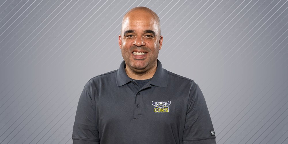 a man smiles while posing for a photo. he is wearing a dark grey shirt with a owl logo on the left side of his chest that says NAIT ooks. the background is blank and light grey