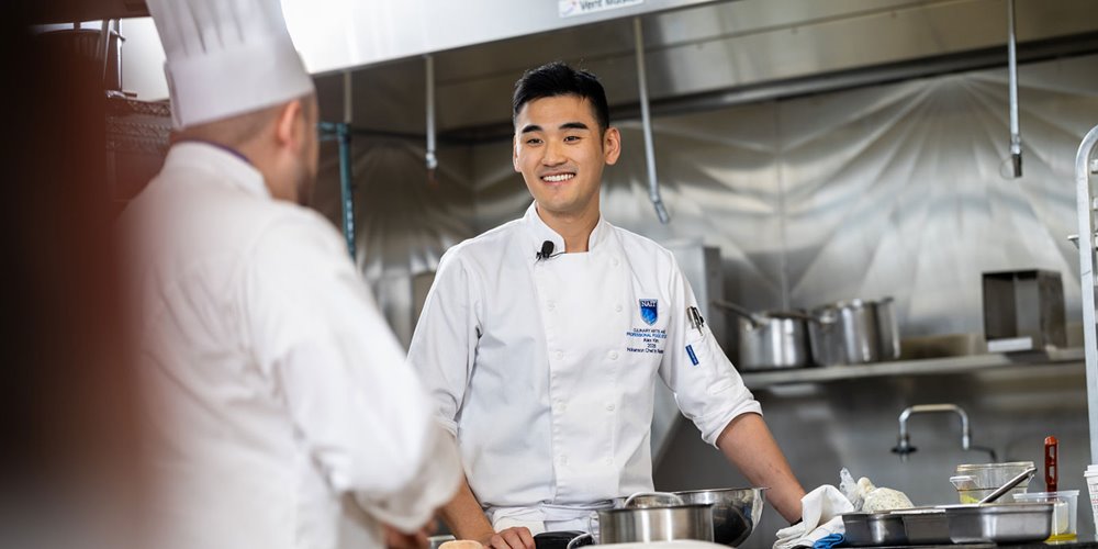 Culinary student in a professional kitchen standing beside a workstation with bowls and utensils, facing an instructor in chef attire.