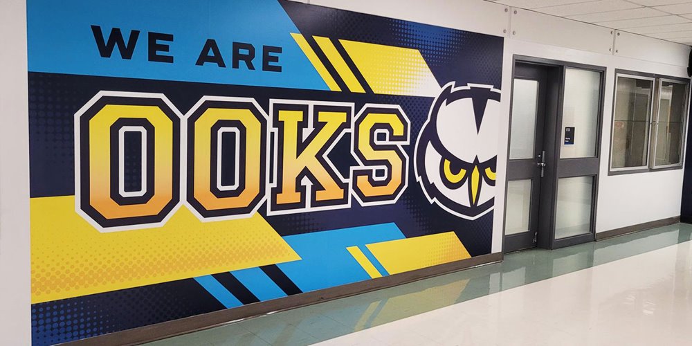 Wall mural in hallway with large text 'WE ARE OOKS' in yellow and black. An illustrated owl face appears next to the word 'OOKS'. Background includes geometric patterns in blue, yellow, and black.