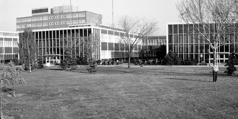 Black-and-white photo of a mid-20th-century institutional building with large glass windows and rectangular sections, surrounded by a grassy lawn with scattered trees.