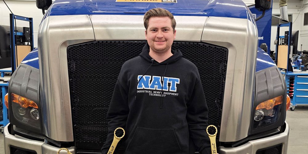 a man in a dark hoodie with the word "NAIT" on it stands in front of a large truck, smiling for a photo. he holds gold-coloured wrenches