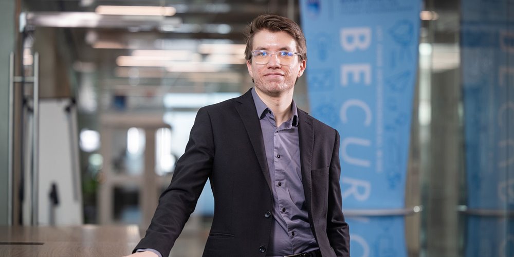 A person wearing a dark suit jacket over a gray dress shirt is standing indoors in a modern office setting, with blue banners in the background displaying text and graphics.