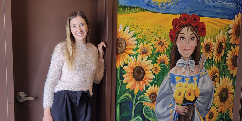 a woman wearing a white sweater and black pants stands in a brown doorway beside a mural of a girl wearing flowers in her hair standing in a field of yellow sunflowers