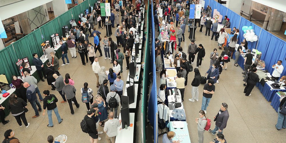 A large indoor event with many people walking around and interacting with various booths. The space is divided by a green curtain on the left and a blue curtain on the right. Each section contains multiple tables or booths with displays, and attendees are gathered around them, engaging in conversations and examining the exhibits.