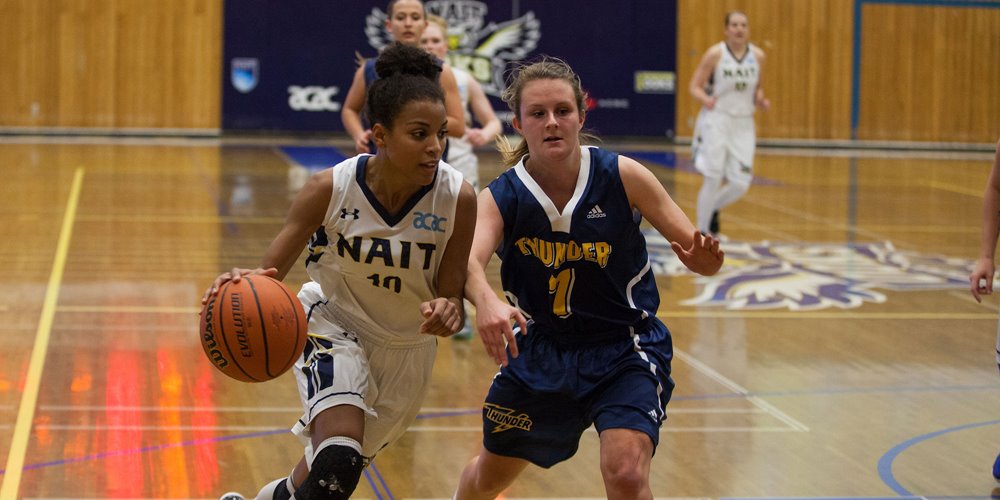 Basketball game between NAIT and Thunder. NAIT player in white jersey (#10) dribbles while Thunder player in blue jersey (#7) defends. Court markings and eagle banner visible in background.