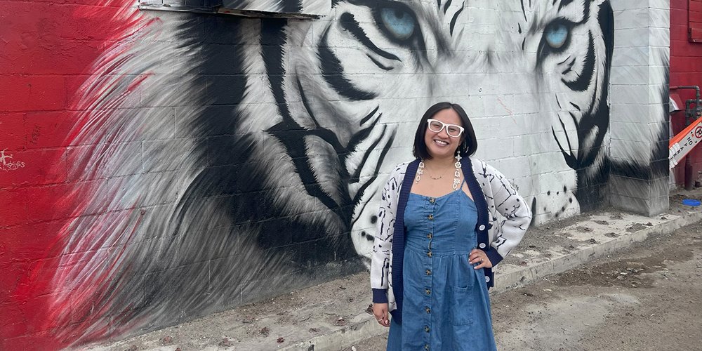 A person in a blue dress and a white cardigan with dark patterns stands with one hand on their hip in front of a mural on a brick wall. The mural features a white tiger with blue eyes, set against a red and black background.